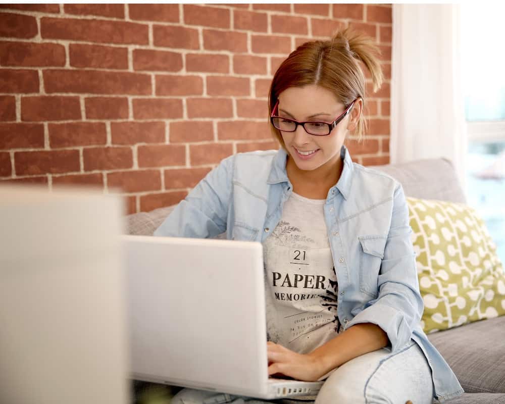 Image of young woman sitting on a couch at home with a laptop- concept of tracking sales at home with a side hustle flipping used clothing.