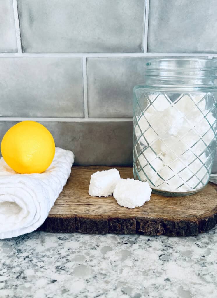 Toilet tabs in a glass jar with two on wooden slab nearby and a lemon on towel.