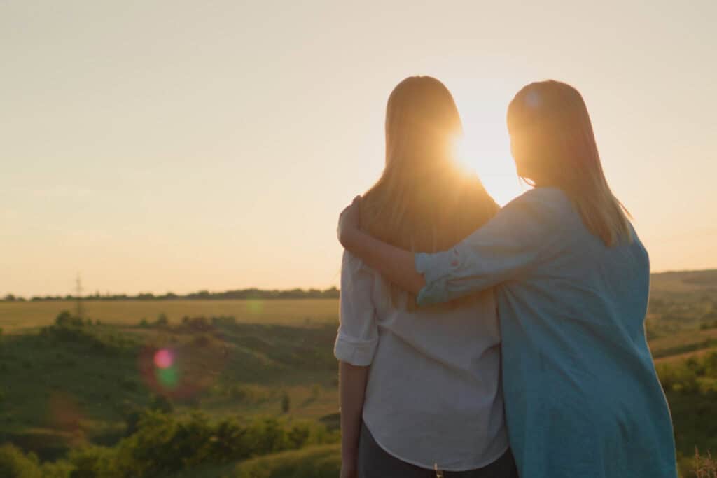 Mom with arm around adult daughter looking at sunset.